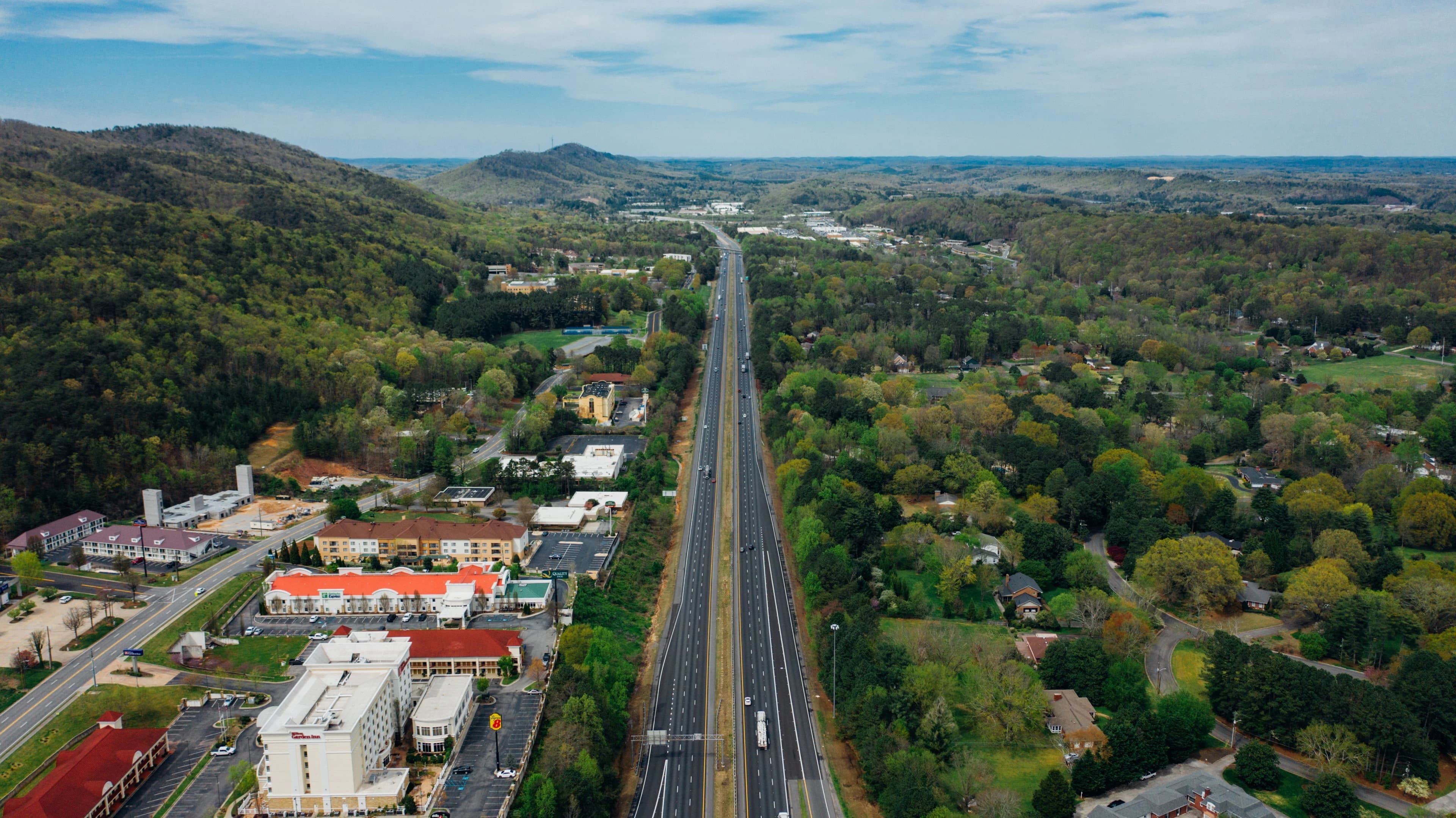 Drone view of narrow asphalt road with driving vehicles surrounded by green forest on hillside and colorful houses in city under spectacular endless sky